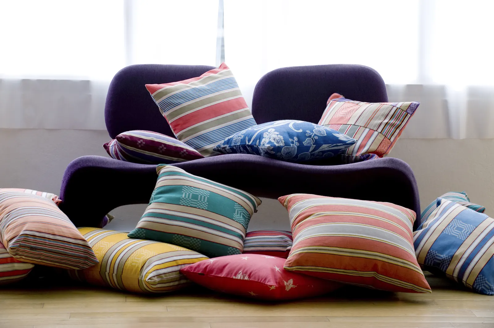 A pile of ticking cushions on a purple armchair by a large window